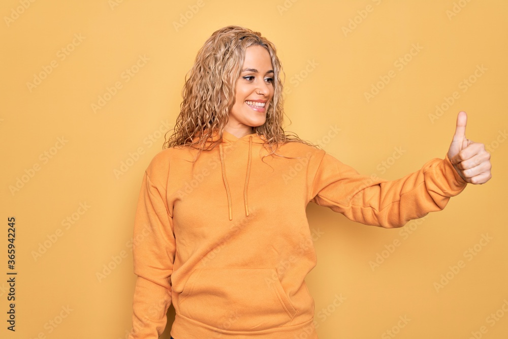 Young beautiful blonde sporty woman wearing casual sweatshirt over yellow background Looking proud, smiling doing thumbs up gesture to the side