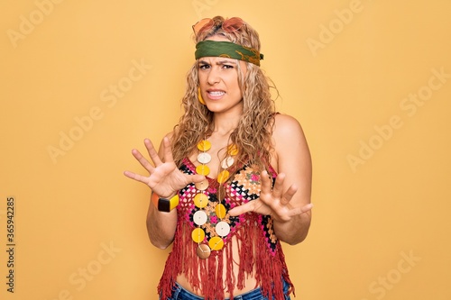 Beautiful blonde hippie woman wearing sunglasses and accessories over yellow background disgusted expression  displeased and fearful doing disgust face because aversion reaction. With hands raised