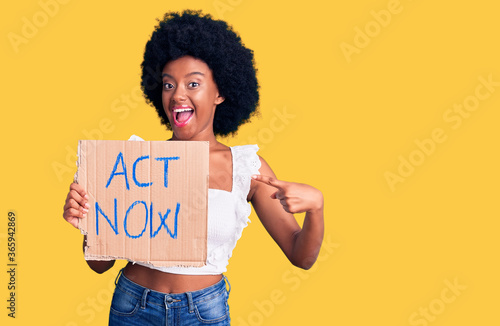 Young african american woman holding act now banner smiling happy pointing with hand and finger photo