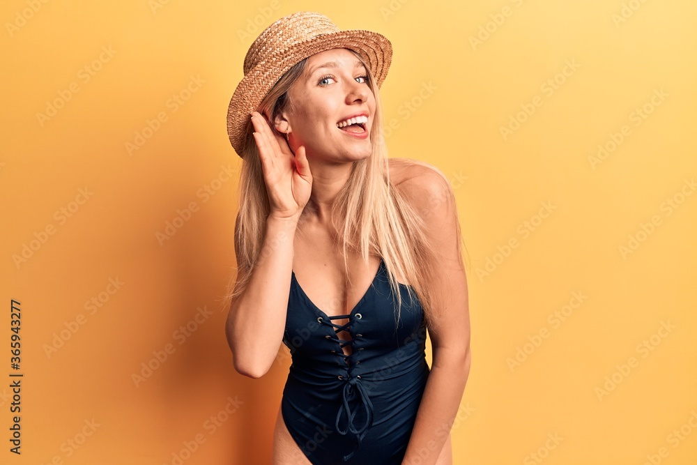 Young beautiful blonde girl wearing swimwear and summer hat smiling with hand over ear listening and hearing to rumor or gossip. deafness concept. Young beautiful blonde girl wearing swimwear and summer hat smiling with hand over ear listening and hearing to rumor or gossip. deafness concept.