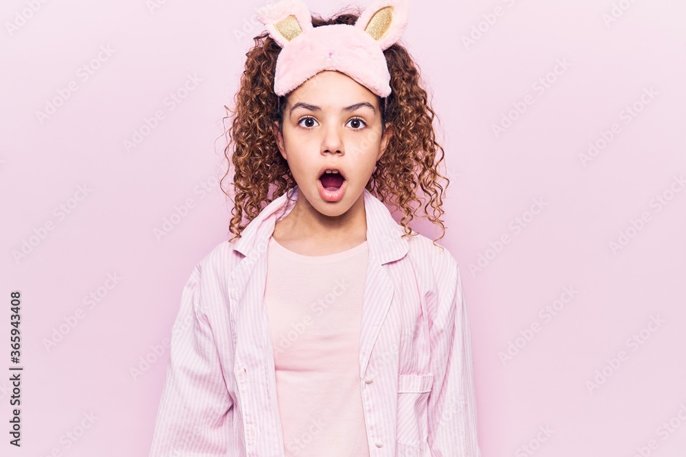 Beautiful kid girl with curly hair wearing sleep mask and pajamas scared and amazed with open mouth for surprise, disbelief face Beautiful kid girl with curly hair wearing sleep mask and pajamas scared and amazed with open mouth for surprise, disbelief face