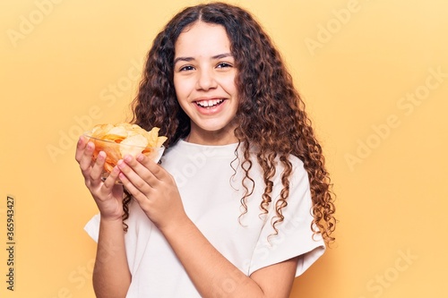 Beautiful kid girl with curly hair holding potato chip looking positive and happy standing and smiling with a confident smile showing teeth