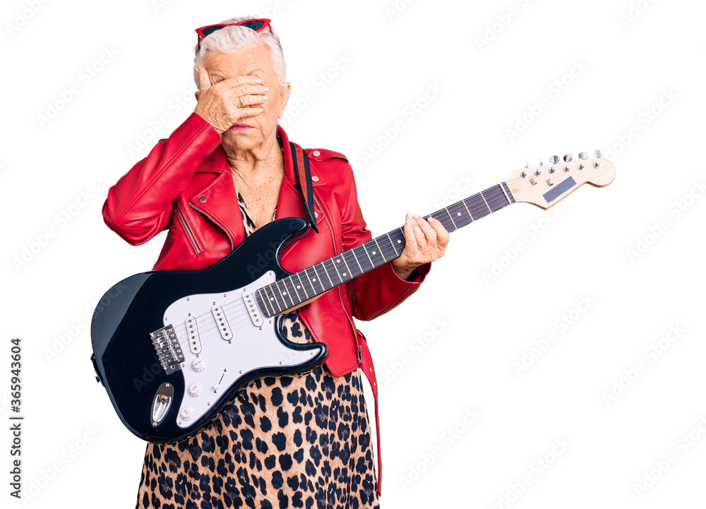 Senior beautiful woman with blue eyes and grey hair wearing a modern look playing electric guitar covering eyes with hand, looking serious and sad. sightless, hiding and rejection concept