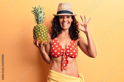 Middle age beautiful woman wearing bikini and summer hat holding pineappel doing ok sign with fingers, smiling friendly gesturing excellent symbol photo