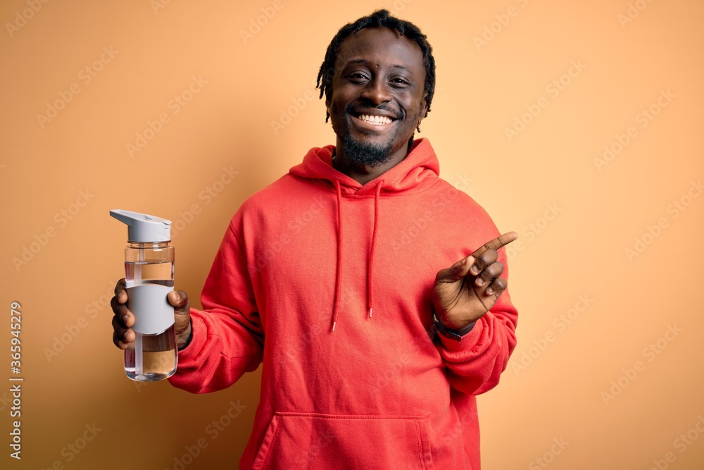 Young african american sporty man wearing sweatshirt with hoodie drinking bottle of water very happy pointing with hand and finger to the side Young african american sporty man wearing sweatshirt with hoodie drinking bottle of water very happy pointing with hand and finger to the side