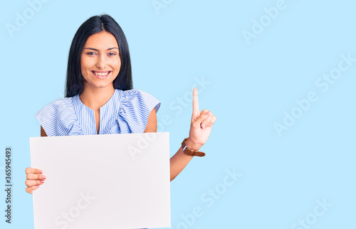 Young beautiful latin girl holding blank empty banner surprised with an idea or question pointing finger with happy face, number one