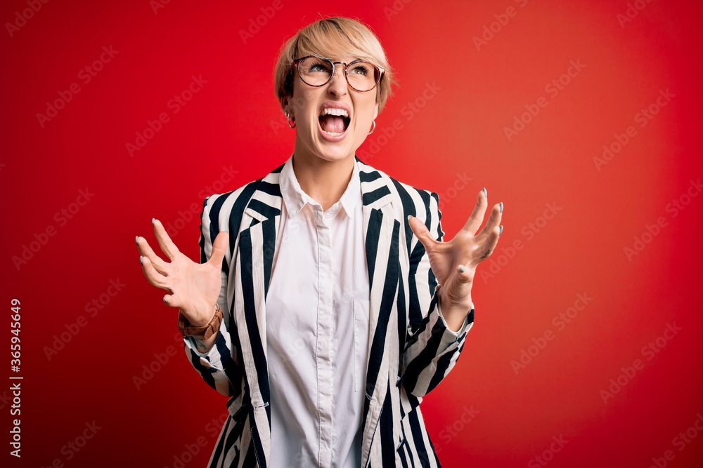 Blonde business woman with short hair wearing glasses and striped jacket over red background crazy and mad shouting and yelling with aggressive expression and arms raised. Frustration concept.