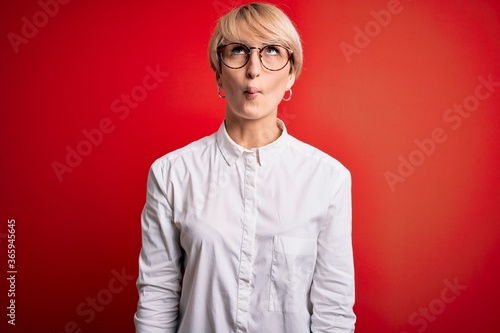 Young blonde business woman with short hair wearing glasses over red background making fish face with lips, crazy and comical gesture. Funny expression. © Krakenimages.com