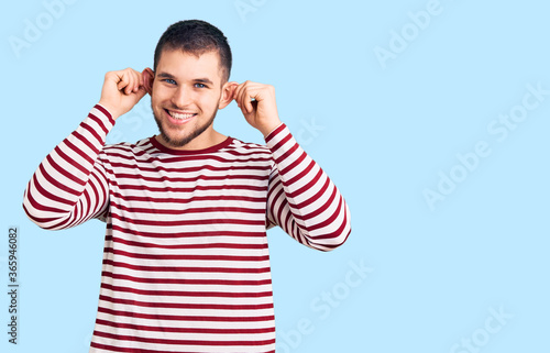 Young handsome man wearing striped sweater smiling pulling ears with fingers, funny gesture. audition problem