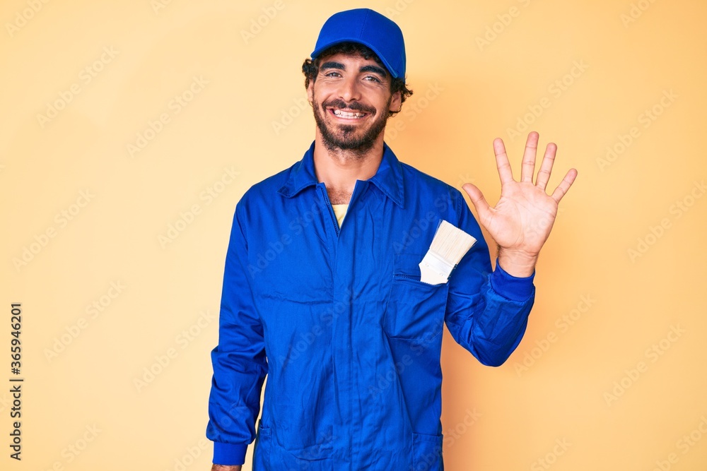 Handsome young man with curly hair and bear wearing builder jumpsuit uniform showing and pointing up with fingers number five while smiling confident and happy.