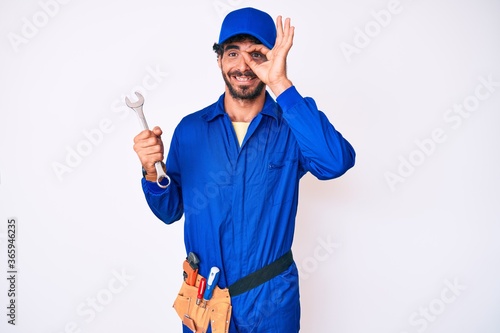 Handsome young man with curly hair and bear wearing builder jumpsuit uniform and holding wrench smiling happy doing ok sign with hand on eye looking through fingers
