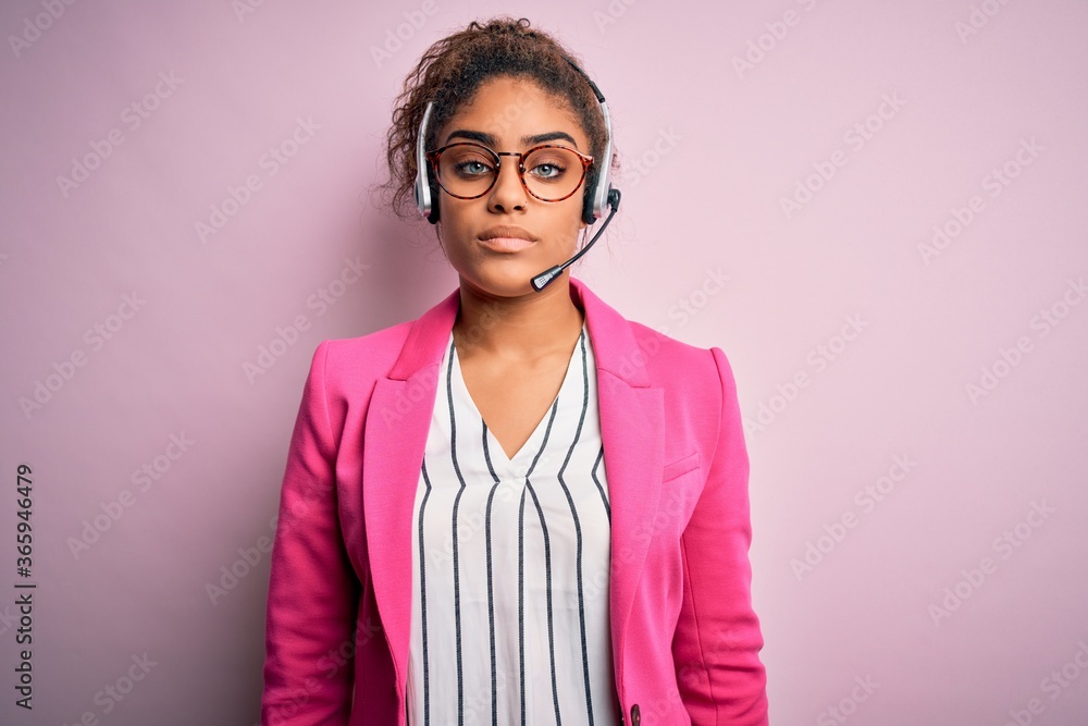 Young african american call center agent girl wearing glasses working using headset with serious expression on face. Simple and natural looking at the camera.