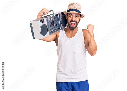 Young hispanic man holding boombox, listening to music screaming proud, celebrating victory and success very excited with raised arms
