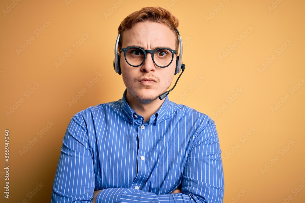 Young handsome redhead call center agent man wearing glasses working using headset skeptic and nervous, disapproving expression on face with crossed arms. Negative person.