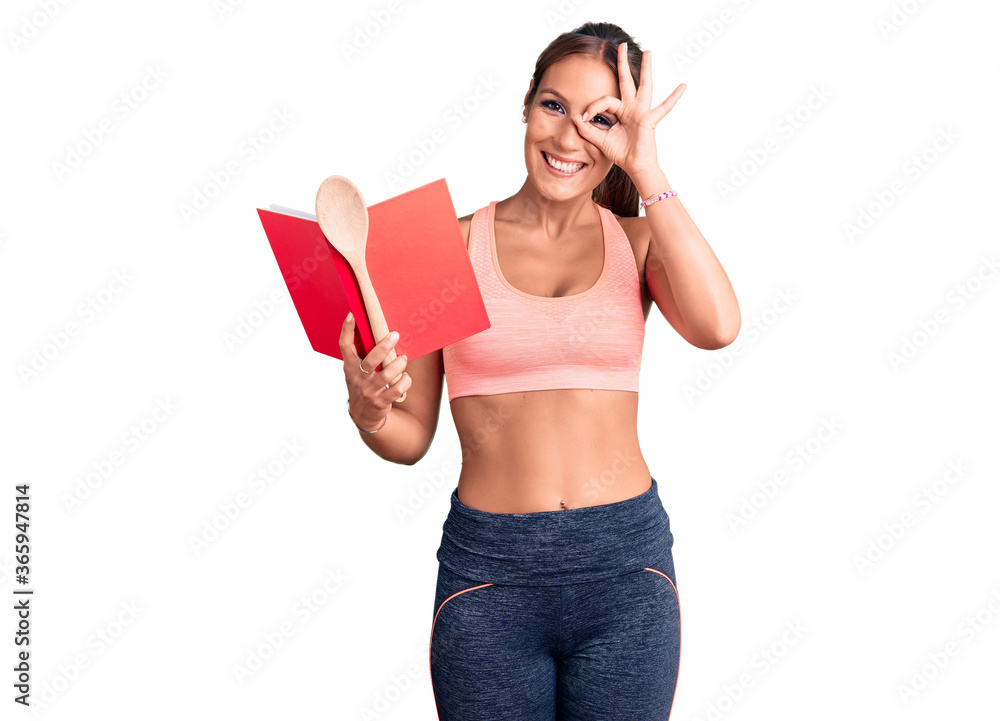 Young beautiful hispanic woman wearing gym clothes reading cooking recipe book holding spoon smiling happy doing ok sign with hand on eye looking through fingers Young beautiful hispanic woman wearing gym clothes reading cooking recipe book holding spoon smiling happy doing ok sign with hand on eye looking through fingers
