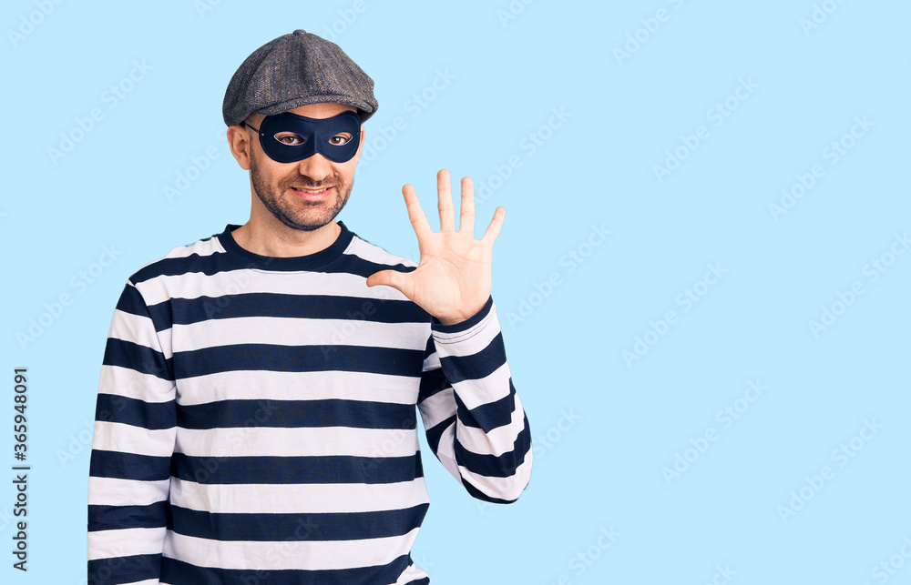 Fototapeta premium Young handsome man wearing burglar mask showing and pointing up with fingers number five while smiling confident and happy.