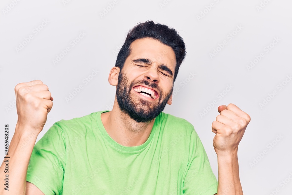 Young handsome man with beard wearing casual t-shirt very happy and excited doing winner gesture with arms raised, smiling and screaming for success. celebration concept. Young handsome man with beard wearing casual t-shirt very happy and excited doing winner gesture with arms raised, smiling and screaming for success. celebration concept.