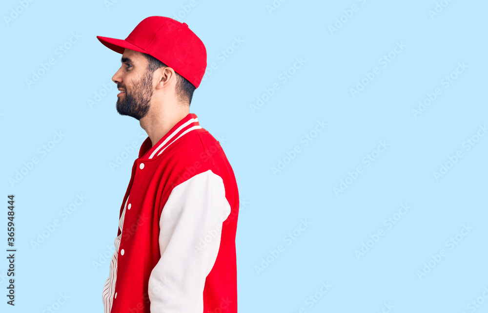 Young handsome man with beard wearing baseball jacket and cap looking to side, relax profile pose with natural face with confident smile.