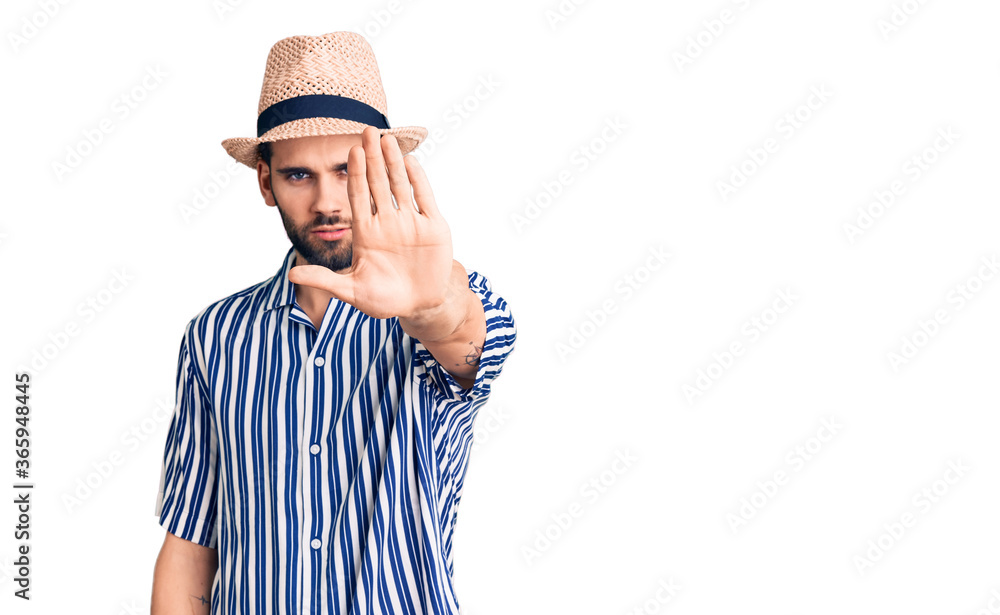 Young handsome man with beard wearing summer hat and striped shirt doing stop sing with palm of the hand. warning expression with negative and serious gesture on the face. Young handsome man with beard wearing summer hat and striped shirt doing stop sing with palm of the hand. warning expression with negative and serious gesture on the face.