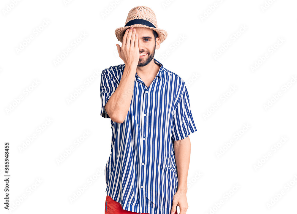 Young handsome man with beard wearing summer hat and striped shirt covering one eye with hand, confident smile on face and surprise emotion. Young handsome man with beard wearing summer hat and striped shirt covering one eye with hand, confident smile on face and surprise emotion.