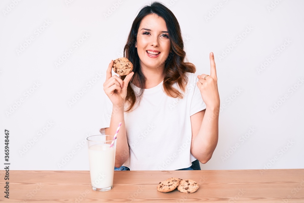 Young beautiful brunette woman drinking a glass of fresh milk with cookies surprised with an idea or question pointing finger with happy face, number one Young beautiful brunette woman drinking a glass of fresh milk with cookies surprised with an idea or question pointing finger with happy face, number one