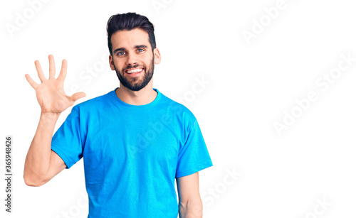 Young handsome man with beard wearing casual t-shirt showing and pointing up with fingers number five while smiling confident and happy.