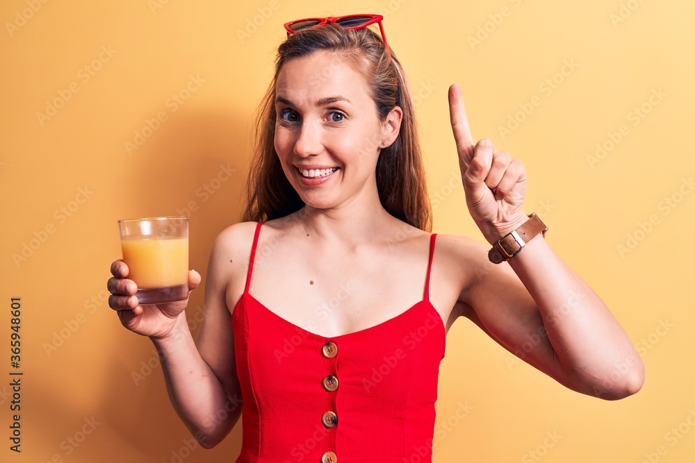Young beautiful blonde woman drinking a glass of orange juice over yellow background smiling with an idea or question pointing finger with happy face, number one Young beautiful blonde woman drinking a glass of orange juice over yellow background smiling with an idea or question pointing finger with happy face, number one
