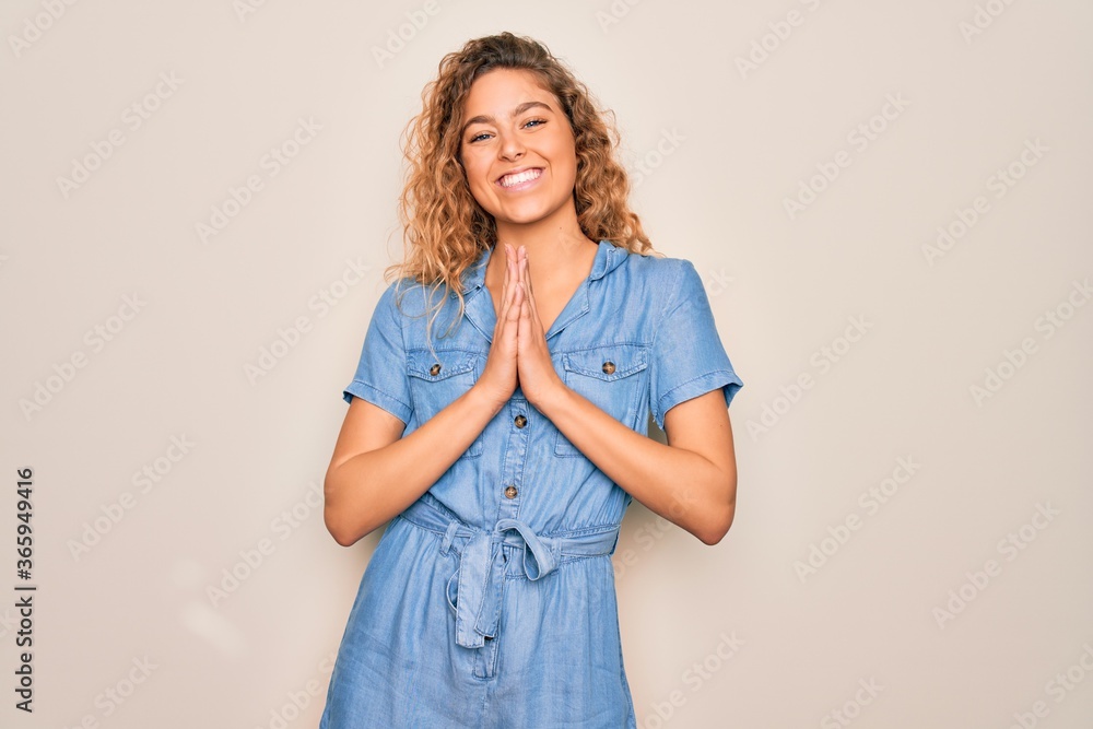 Φωτογραφία Young beautiful woman with blue eyes wearing casual denim dress over white background praying with hands together asking for forgiveness smiling confident
