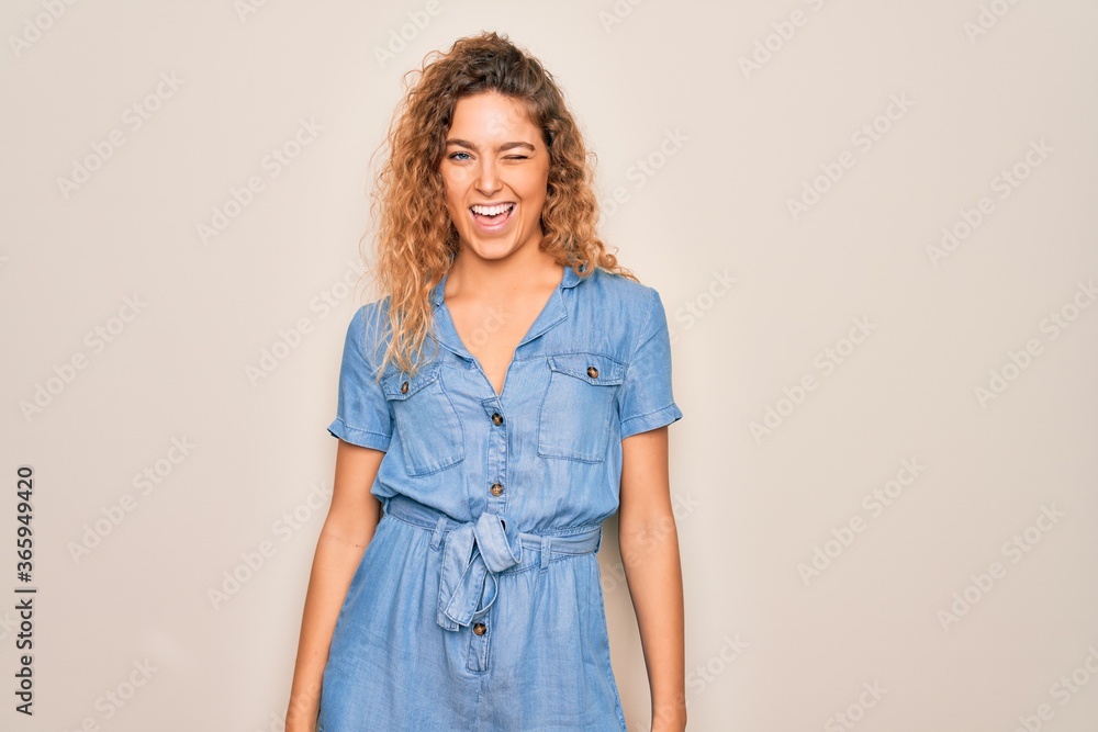 Young beautiful woman with blue eyes wearing casual denim dress over white background winking looking at the camera with sexy expression, cheerful and happy face. Young beautiful woman with blue eyes wearing casual denim dress over white background winking looking at the camera with sexy expression, cheerful and happy face.