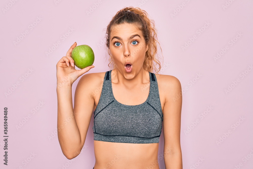 Young beautiful blonde sporty woman with blue eyes holding healthy green apple fruit scared in shock with a surprise face, afraid and excited with fear expression Young beautiful blonde sporty woman with blue eyes holding healthy green apple fruit scared in shock with a surprise face, afraid and excited with fear expression