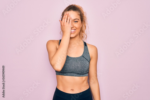 Young beautiful blonde sportswoman with blue eyes doing exercise wearing sportswear covering one eye with hand, confident smile on face and surprise emotion.