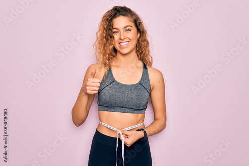 Young blonde sporty woman with blue eyes controlling weight using tape measure on waist happy with big smile doing ok sign, thumb up with fingers, excellent sign © Krakenimages.com