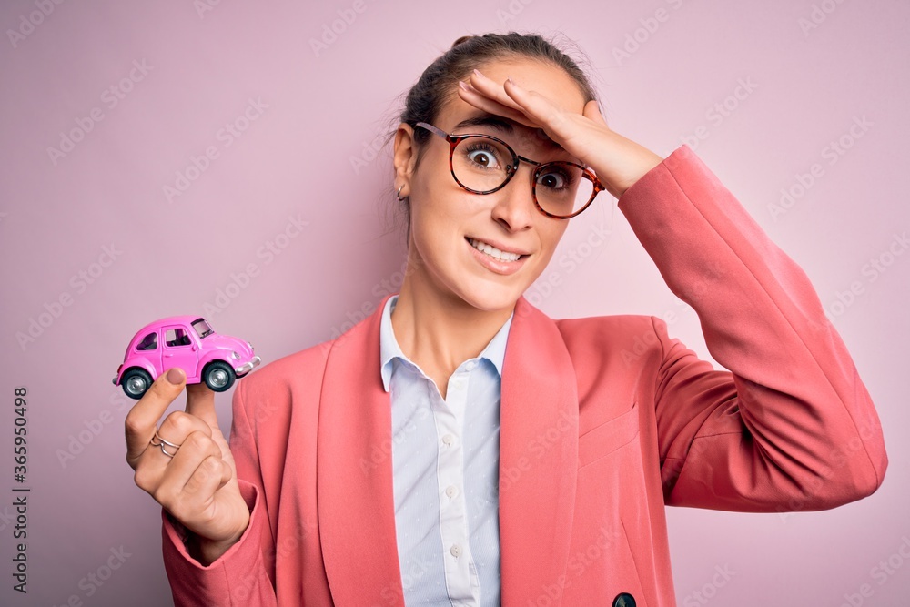 Young beautiful businesswoman wearing glasses holding pink car toy over isolated background stressed with hand on head, shocked with shame and surprise face, angry and frustrated. Fear and upset Young beautiful businesswoman wearing glasses holding pink car toy over isolated background stressed with hand on head, shocked with shame and surprise face, angry and frustrated. Fear and upset