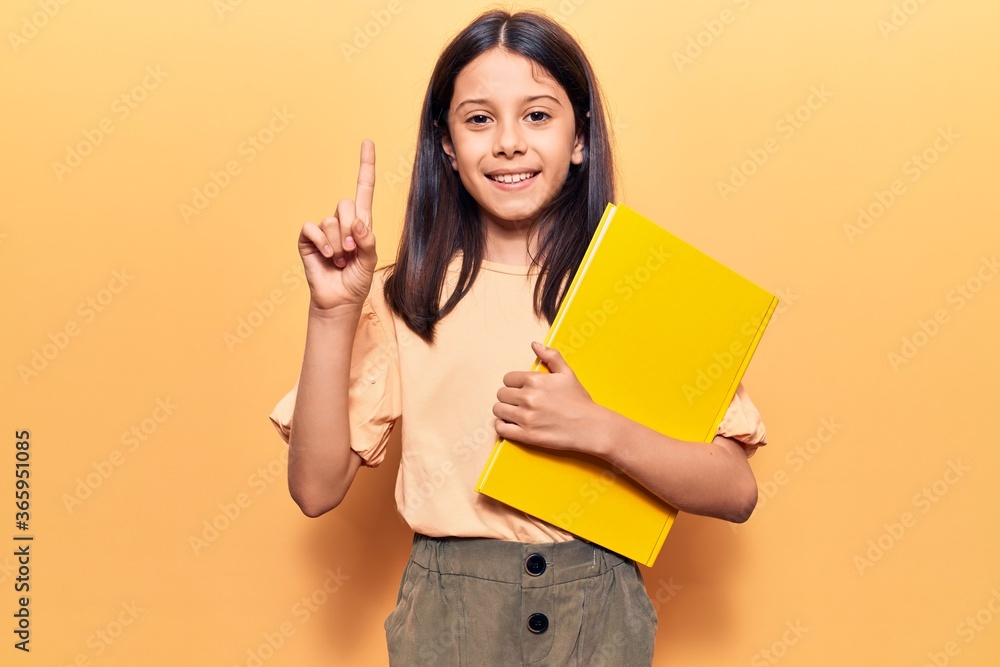 Beautiful child girl holding book smiling with an idea or question pointing finger with happy face, number one Beautiful child girl holding book smiling with an idea or question pointing finger with happy face, number one