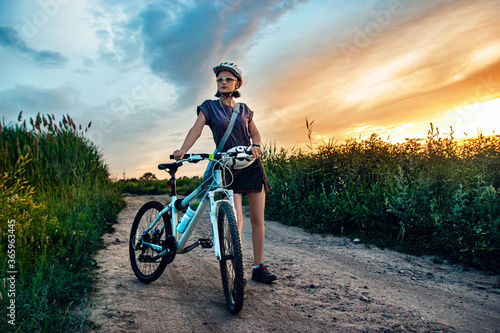 Young woman and mountain bike stands in the rural green field at summer evening. Adventure summer leisure time.