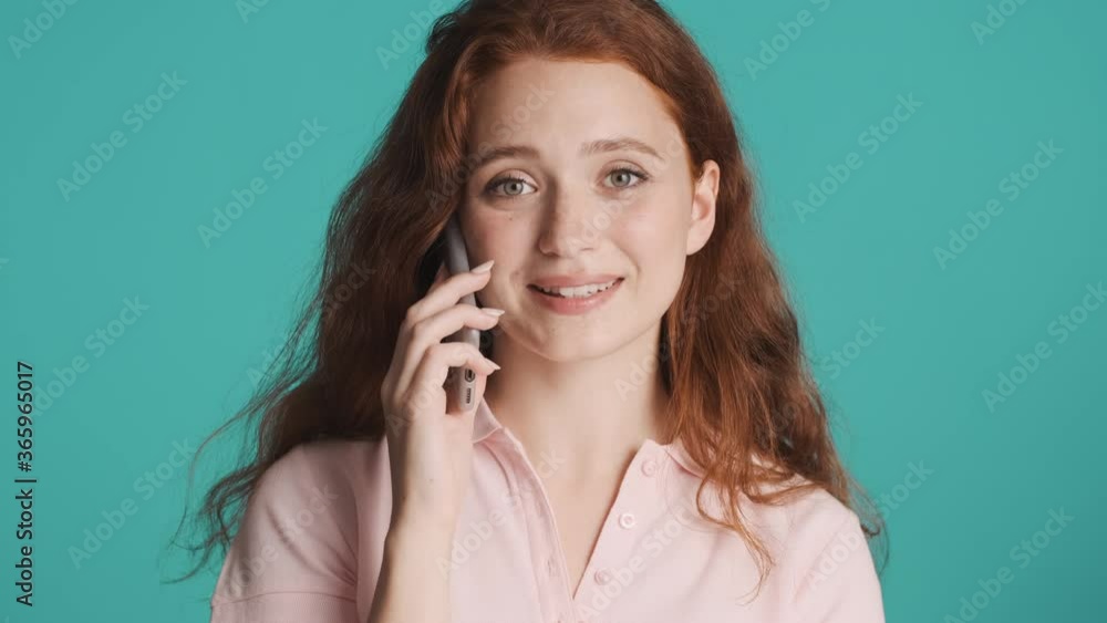 Beautiful charming redhead girl happily talking on phone and smiling in camera over colorful background Beautiful charming redhead girl happily talking on phone and smiling in camera over colorful background