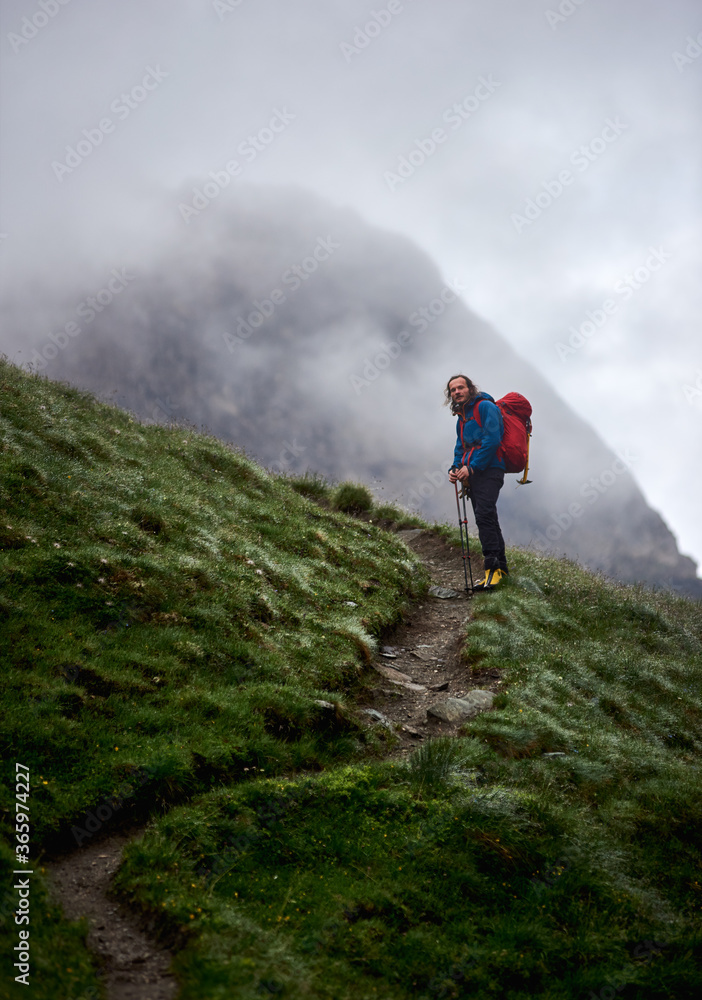 Man traveler with backpack standing on grassy hillside path with foggy mountain on background. Handsome tourist looking at camera while hiking in mountains. Concept of traveling and climbing.