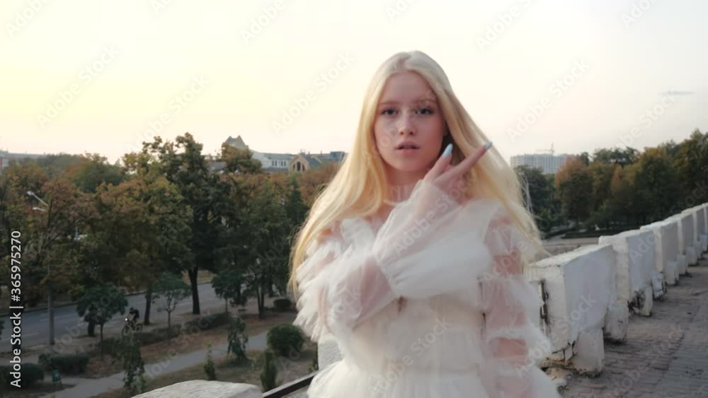 Accelerated cinematic close-up shot charming cute blonde girl with long silky hair and stylish white dress walking on observation deck against background of spring trees in center of Kharkov, Ukraine. Accelerated cinematic close-up shot charming cute blonde girl with long silky hair and stylish white dress walking on observation deck against background of spring trees in center of Kharkov, Ukraine.