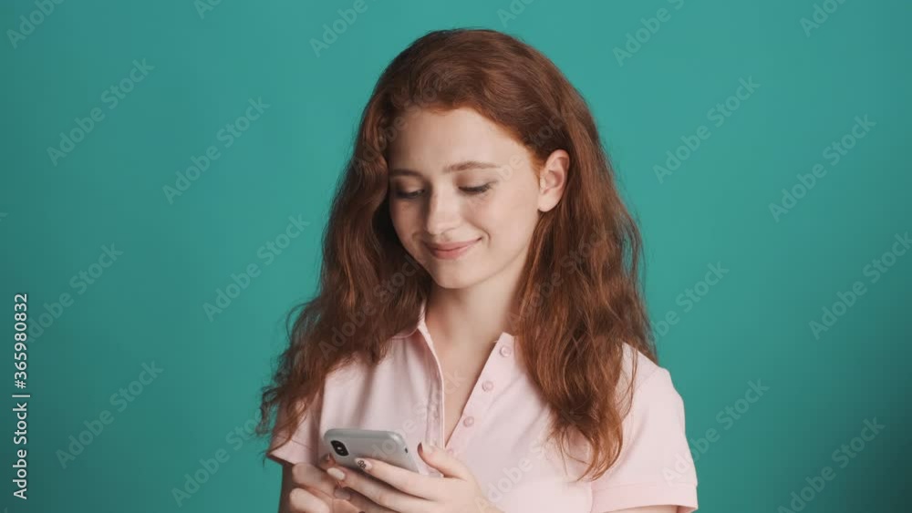 Pretty casual redhead girl chatting with friends on smartphone and smiling over colorful background Pretty casual redhead girl chatting with friends on smartphone and smiling over colorful background