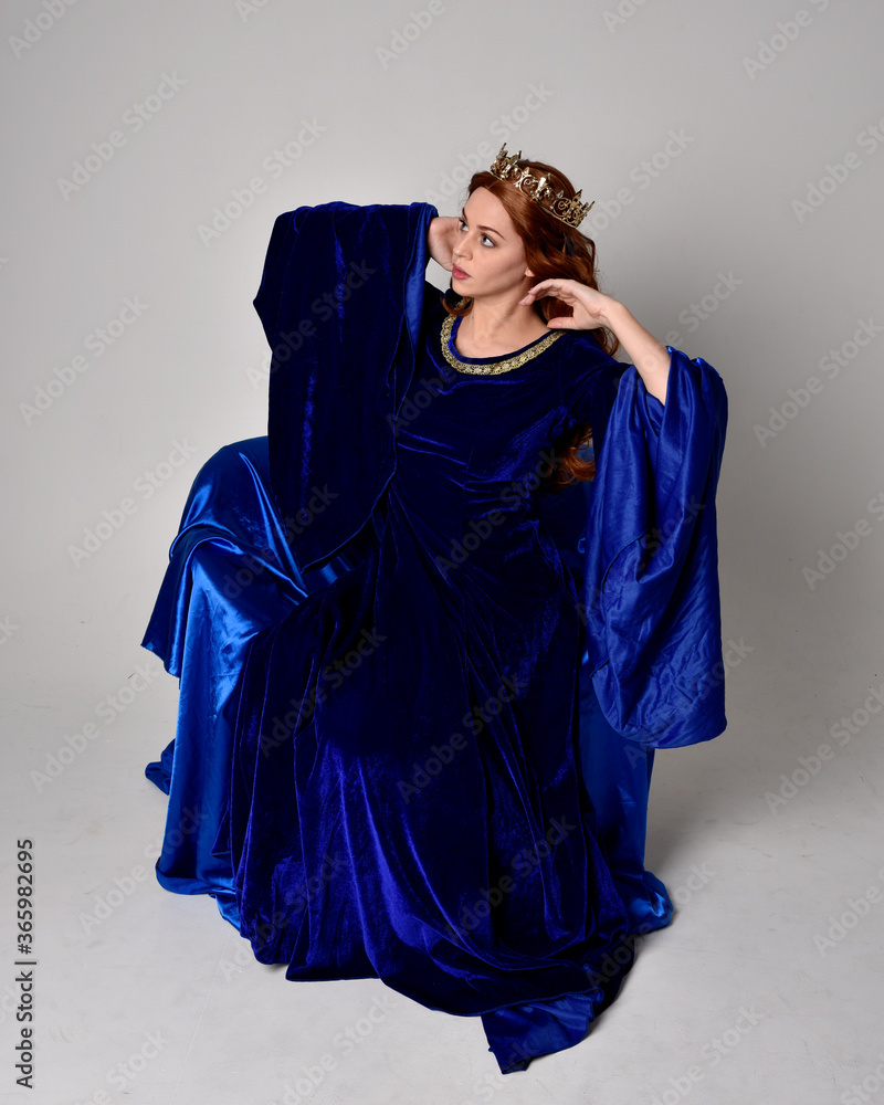 Full length portrait of girl wearing long blue velvet gown with golden crown. sitting pose, isolated against a studio background. Full length portrait of girl wearing long blue velvet gown with golden crown. sitting pose, isolated against a studio background.