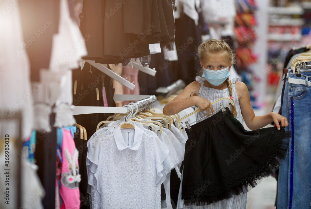 Little girl in a medical mask chooses a skirt to school. School uniform store. Preparing for school after quarantine Covid - 19.