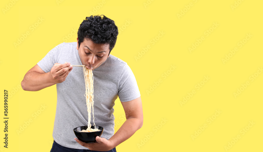 Young handsome trendy Asian man eating yummy hot and spicy instant noodle using chopsticks and bowl isolated on yellow background in studio With copy space. Young handsome trendy Asian man eating yummy hot and spicy instant noodle using chopsticks and bowl isolated on yellow background in studio With copy space.