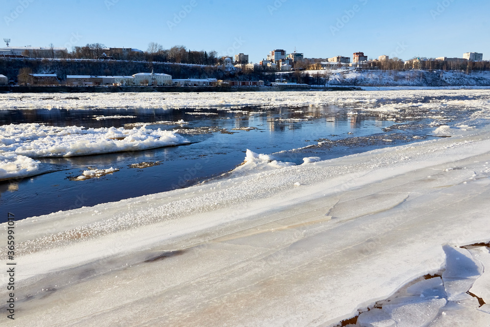 Ice drift on a river with blue high water and big water, white snow broken ice full of hummocks in it and city with houses at a background in sunny spring day.