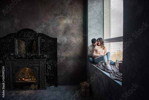 Happy couple in love in winter sweaters and jeans, sit on a wide windowsill by the window, hug. A young man kisses a beautiful curly brown-haired girl on the cheek. Date by the fireplace in cozy home