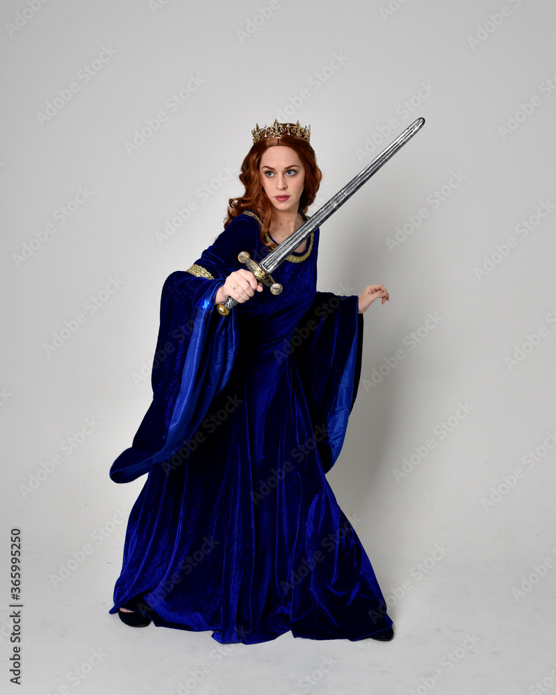 Full length portrait of girl wearing long blue velvet gown with golden crown, holding a sword. standing pose isolated against a studio background. Full length portrait of girl wearing long blue velvet gown with golden crown, holding a sword. standing pose isolated against a studio background.