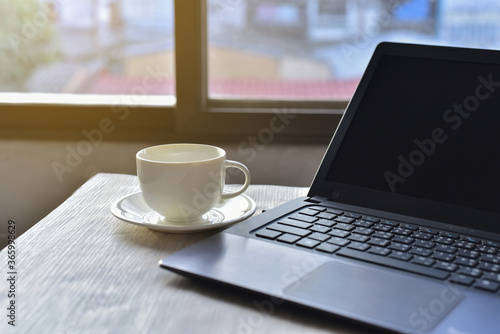 white coffee cup beside the black laptop againt the sunrise from the windows