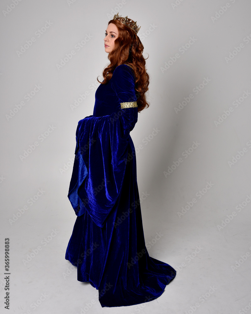 Full length portrait of girl wearing long blue velvet gown with golden crown. standing pose with back to the camera, isolated against a studio background. Full length portrait of girl wearing long blue velvet gown with golden crown. standing pose with back to the camera, isolated against a studio background.