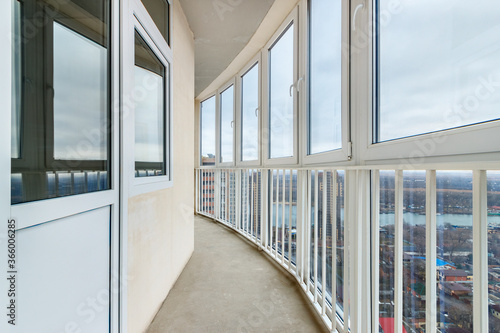 A curved balcony with the panoramic windows on the high floor of a new residential building. A loggia with the white plastered walls without decoration