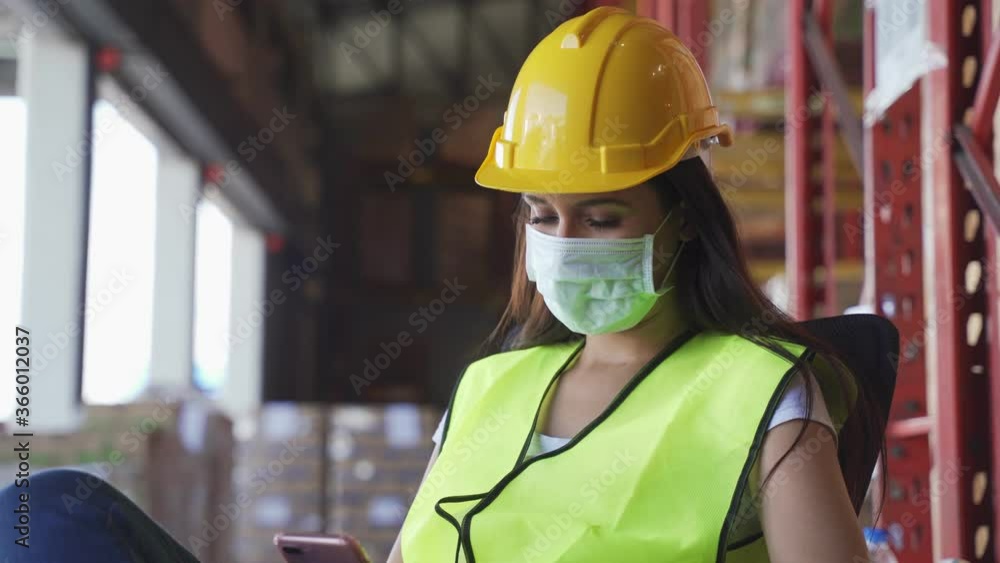 Young engineer worker wearing face mask and vest working in shipping logistic warehouse factory during Covid-19 pandemic. Girl sitting on chair take a rest using mobile phone feeling relax from work. Young engineer worker wearing face mask and vest working in shipping logistic warehouse factory during Covid-19 pandemic. Girl sitting on chair take a rest using mobile phone feeling relax from work.