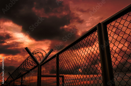 Security fence of military zone or private area fence with red sky and dark clouds. Barbed wire security fence with stormy sky. Barrier border. Boundary security wall. Dramatic and horror sky. © Artinun